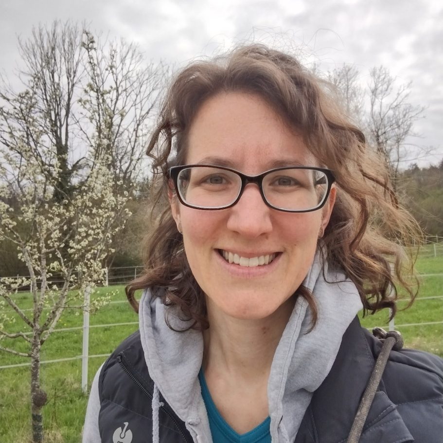 Yvi Schuler - Dein ANIfit Berater Frau mit Locken und Brille lächelt vor einer Landschaft mit Bäumen und Wiese.