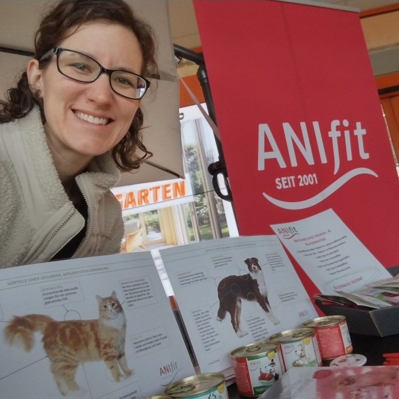 Lächelnde Frau mit Brille steht vor einem Stand für Tiernahrung und -produkte.