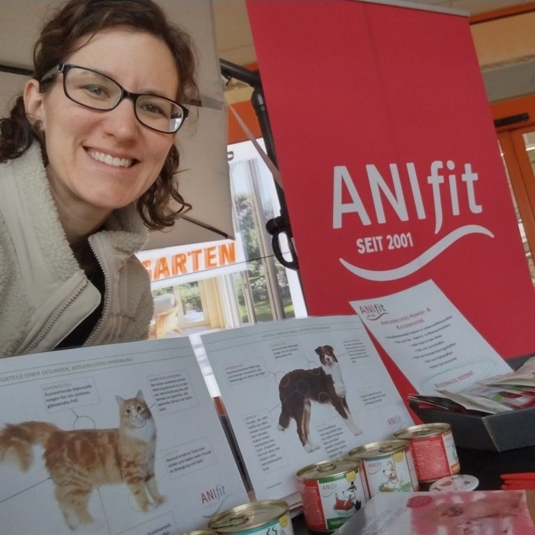 Kostenlose Futterberatung mit ANIfit im Zollernalbkreis Eine lächelnde Frau steht am ANIfit-Stand mit Tierfutter und Informationsblättern.