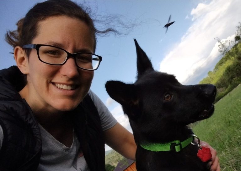 Frau mit Brille lächelt mit ihrem Hund auf einer Wiese im Freien.