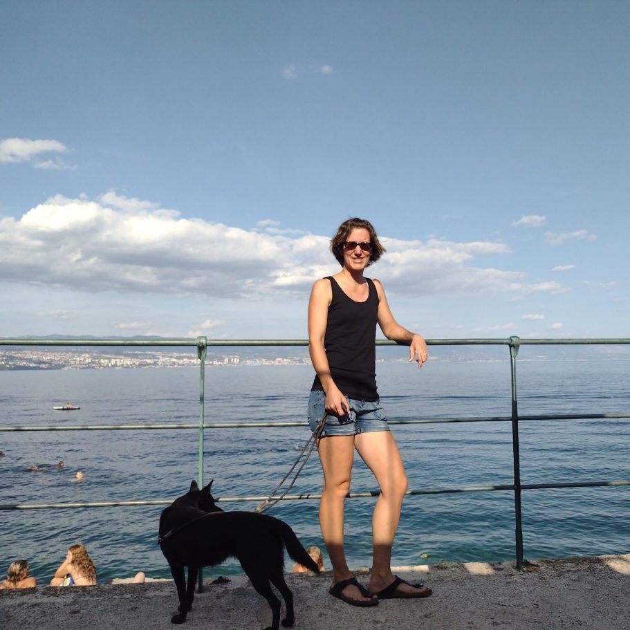 Frau in Shorts und T-Shirt steht neben einem schwarzen Hund am Wasser.