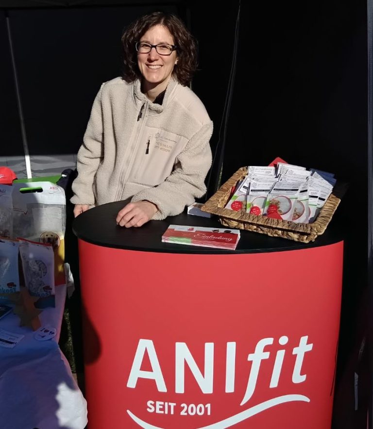 Frau steht an einem Tisch mit ANIfit-Produkten und lächelt freundlich.