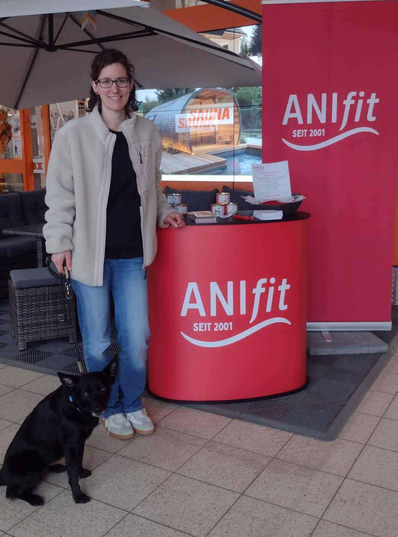 Frau steht neben einem Tisch mit ANIfit-Logo, schwarzer Hund sitzt davor.