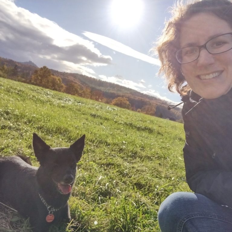 Yvis Futterberatung | Dein ANIfit Berater Eine Frau sitzt im Gras neben einem schwarzen Hund, sonniger Tag mit blauen Himmel.